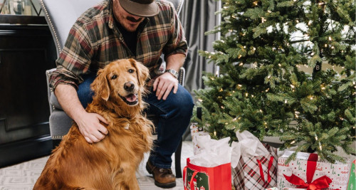 A man and his Golden Retriever opening gifts by a Christmas tree