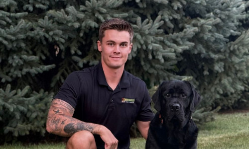 Dog trainer kneeling on grass beside a black Labrador retriever