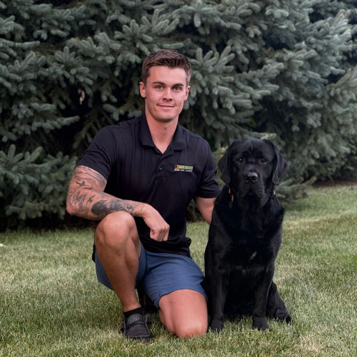 Dog trainer kneeling on grass beside a black Labrador retriever